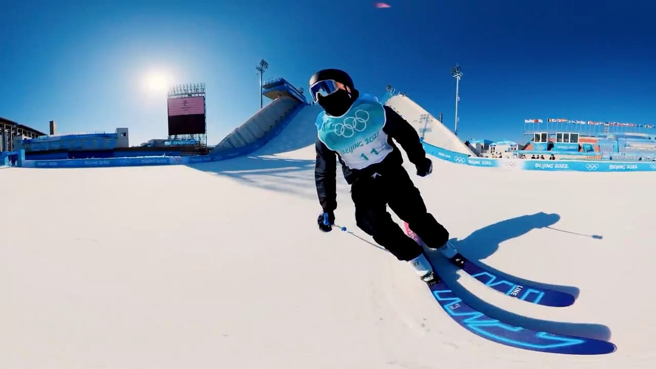 A freestyle skier in a Beijing 2022 competition bib carving across the landing at Big Air Shougang, skis sweeping through frame, the take-off ramp and floodlights behind against a deep blue sky — captured on the 360° camera whose lens curves the horizon.